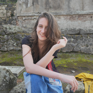 Mara Sergienko sitting outside, with her hair swept slightly over her face and she is holding sunglasses.