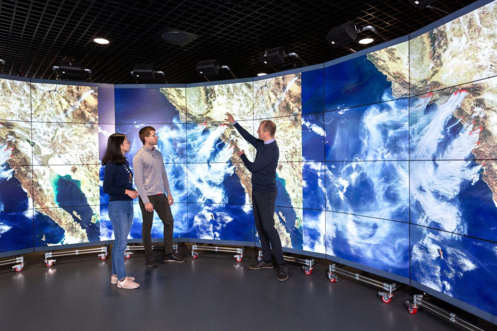 Three people stand in front of screens displaying a satellite image of the Earth.