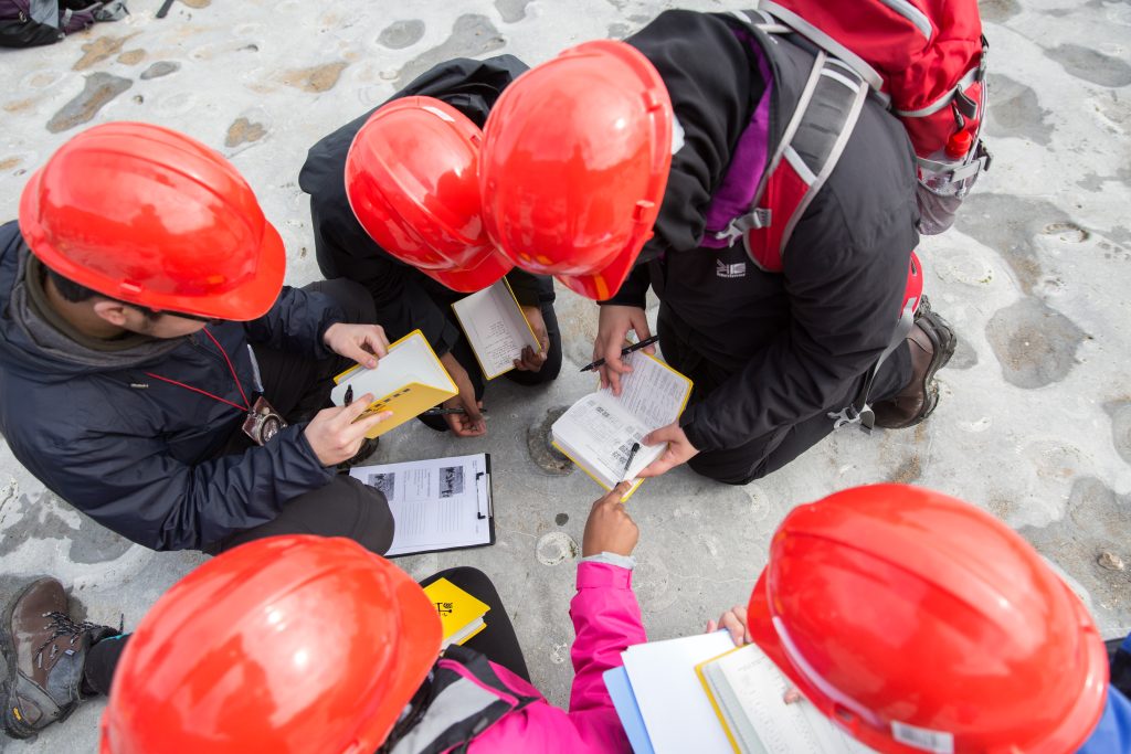 Students wearing red helmets gather in a circle around their notebooks during fieldwork.