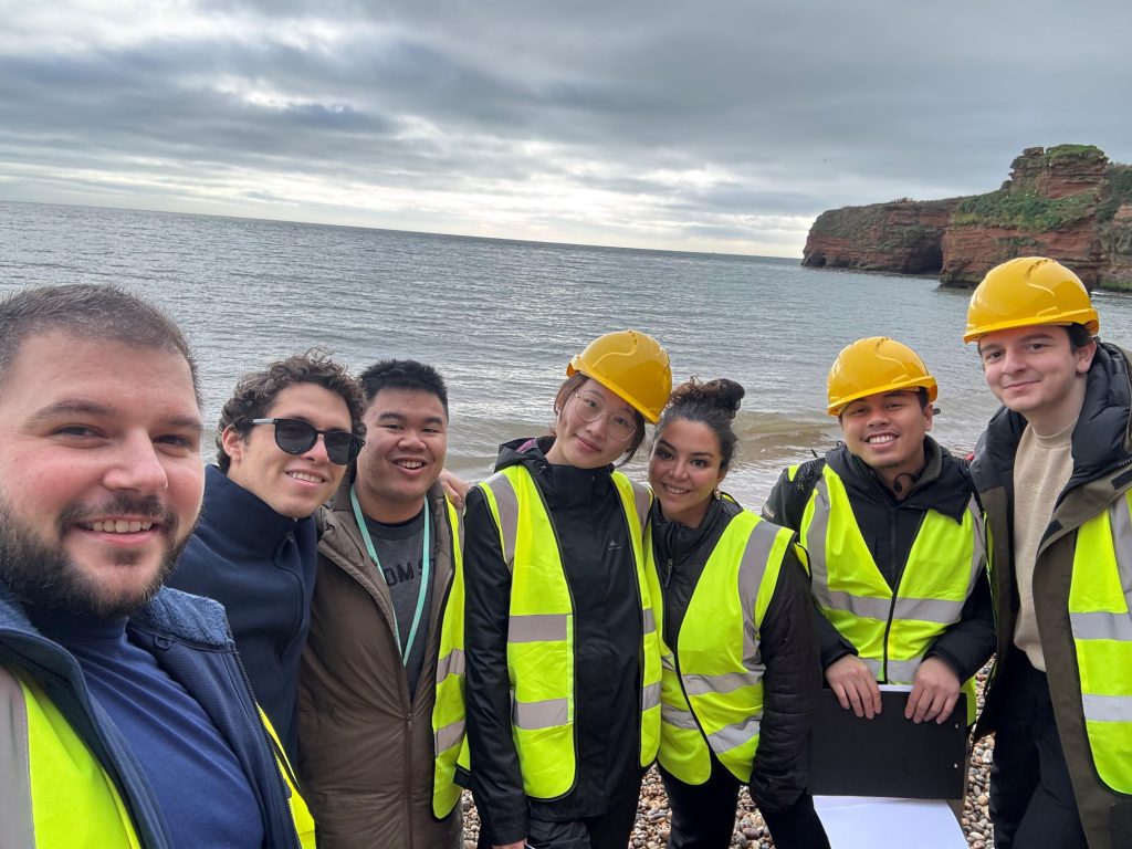 A group of students during a Metals and Energy Finance MSc field trip stand in front of the sea, wearing helmets. 