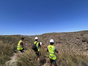 3 students wearing hi-vis and helmets looking across a valley towards an ancient reef complex. They are drawing a sketch of the outcrop into their yellow notebooks