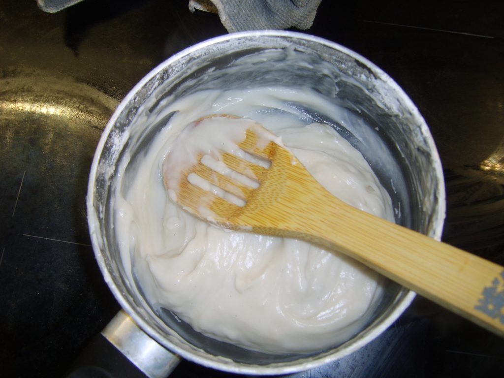 A photo of a saucepan on a stove. A wooden spoon is being used to stir the thick white mixture inside.