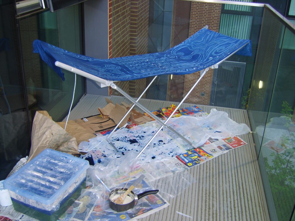 Photo of a washing airer on a balcony surrounded by newspaper and dyeing equipment. On the airer is a long piece of blue fabric.