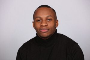 Portrait of De-Shaine Murray. De-Shaine is sat in front of a white backdrop, he is wearing a black turtle neck