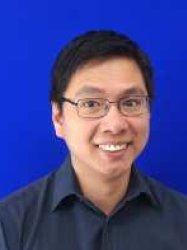 Portrait of Joel Eustaquio. Joel stands smiling in front of a blue backdrop wearing a navy blue short and rectangular glasses