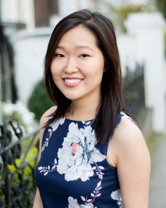 Portrait of Julia Sun. Julia stands outdoors smiling. She is wearing a dress that is blue and has white floral details 