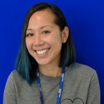 Portrait of Julie Hoang. Julie is sat smiling in front of a blue backdrop. She is wearing a grey jumper over a white shirt and black jeans.