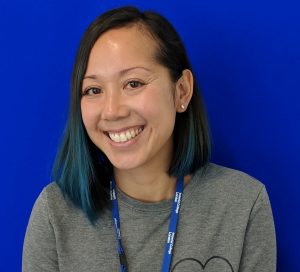 Portrait of Julie Hoang. Julie is sat smiling in front of a blue backdrop. She is wearing a grey jumper over a white shirt and black jeans.