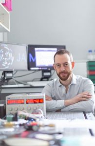 Joseph is at a desk with his arms crossed. An early prototype of the JAMVENT ventilator is on the desk and PC monitors can be seen in the background