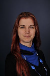 Portrait of Edit Toth. Edit is standing in front of a blue backdrop. She is smiling and wearing a purple jumper with a black cardigan