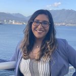 Portrait of Aditi Sitolay standing outdoors by a body of water, smiling at the camera, with mountains and a harbour visible in the background.