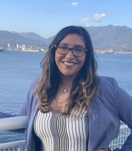 Portrait of Aditi Sitolay standing outdoors by a body of water, smiling at the camera, with mountains and a harbour visible in the background.