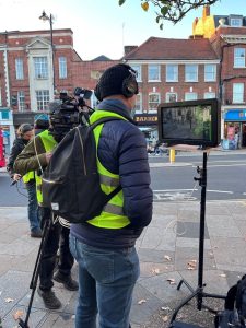 A man stands in a street, wearing a hat, headphones and backpack, looking at a screen on a tripod. A person with a video camera is visible in the background.