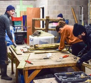 Three people in a workshop with planks for wood which are being hammered and sawn.