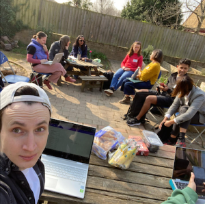 A group of people sat outside with a person in the foreground taking a selfie.