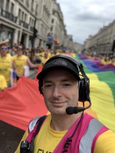 A man wearing a headset with a microphone, a baseball cap and a high vis jacket, with a rainbow flag in the background