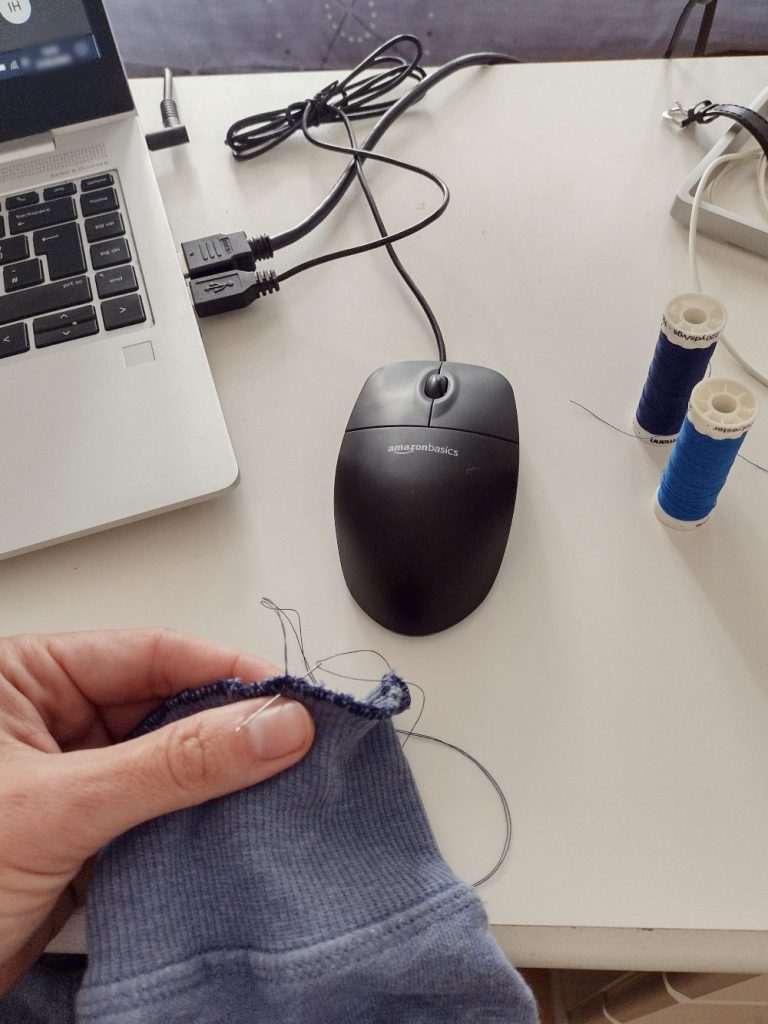 Desk surface with bobbins, mouse and laptop. A feminine hand is holding a blue sleeve with needle and thread attached