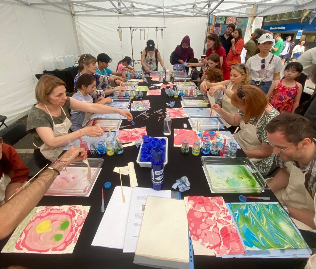 Group of people of all ages sitting round a table in a tent. The table is covered art materials. Each participant is creating a marbled image. 