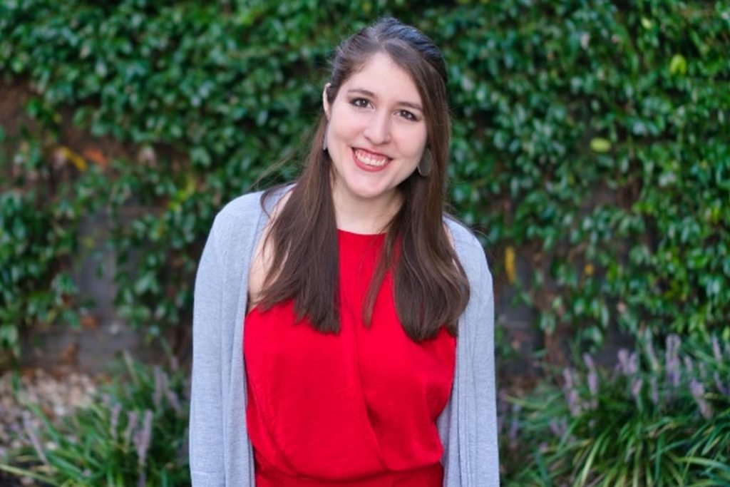 Javiera Perez, wearing a red dress, smiling at the camera and standing against an ivy covered wall