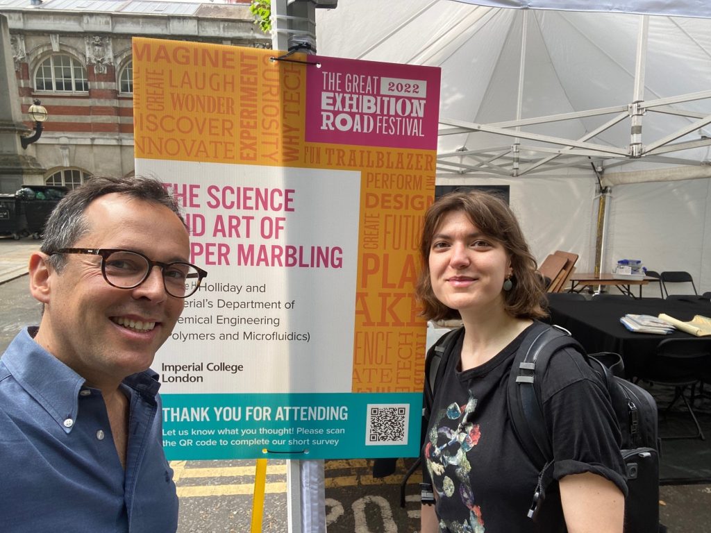 Joao Cabral and Liva Donina grinning in front of the poster for the marbling activity at the 2022 Great Exhibition Road Festival