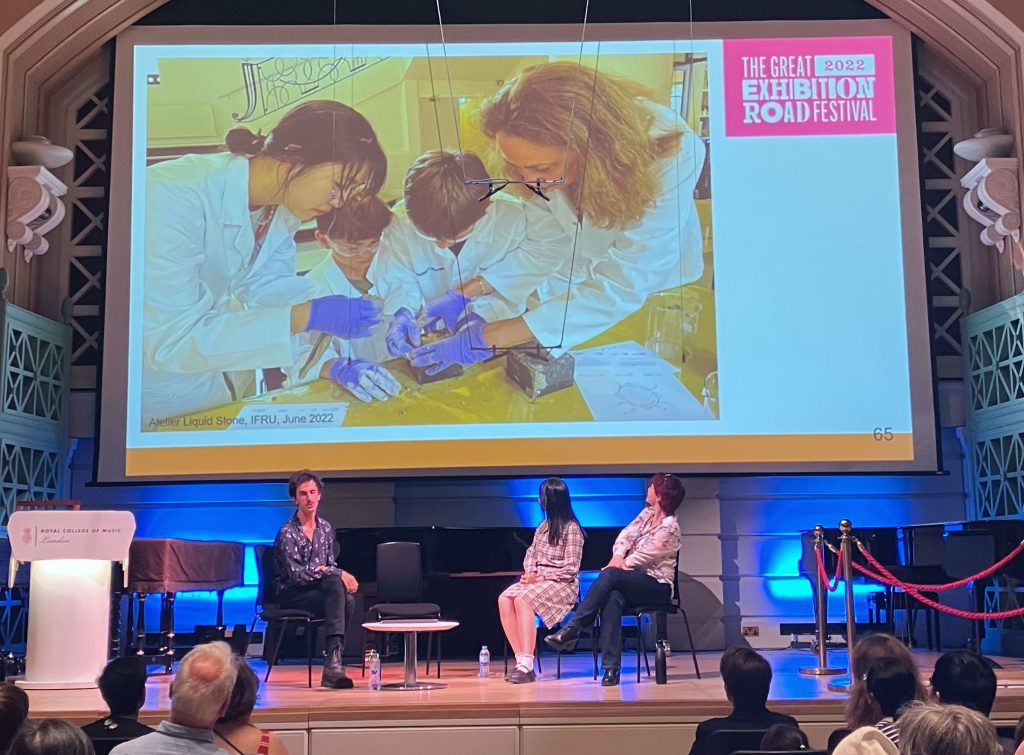 Clement Périssé, Chen Gong and Mary Ryan on stage at the Royal Academy of Music. Above their heads is a projector screen showing a photograph of Chen and another teacher working with 2 children to make a polyhedral 'moss ball'