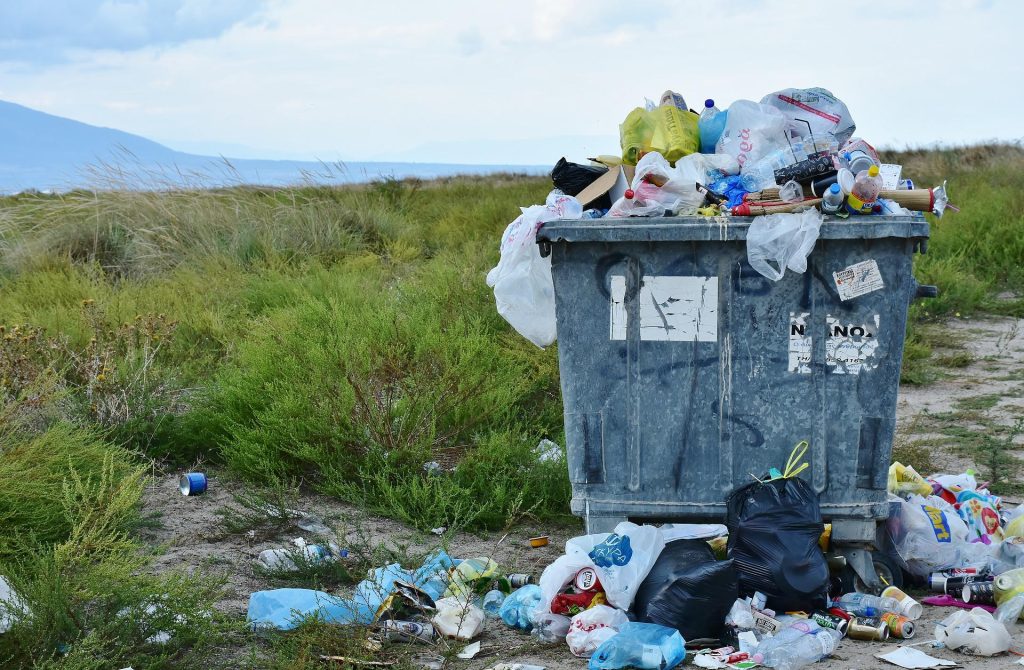 A very full rubbish container, with bags of rubbish spilled around it, at the edge of a beach