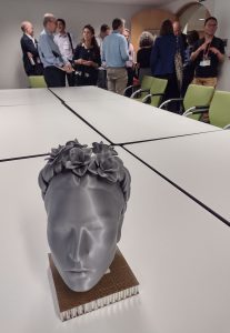 A 3D printed head of Frida Kahlo stands on a table in a meeting room. In the background a group of researchers are standing and talking.