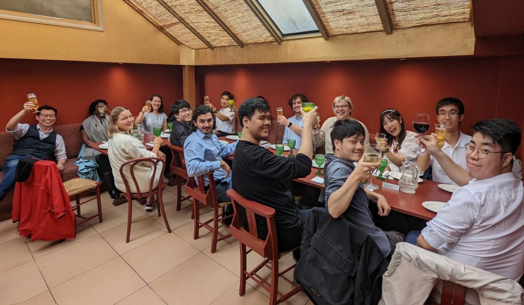 A group of students and staff sitting around restaurant tables raise a glass to the camera in celebration