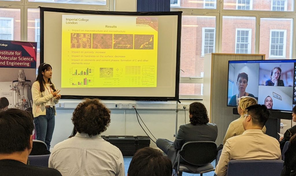 A female student stands in front of an audience with her slides displayed behind her; on the right is a screen displaying the faces of a number of people observing the meeting remotely