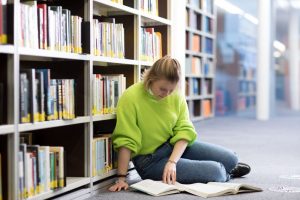 A woman student sitting on the floor in front of library shelves, reading from two open books. 