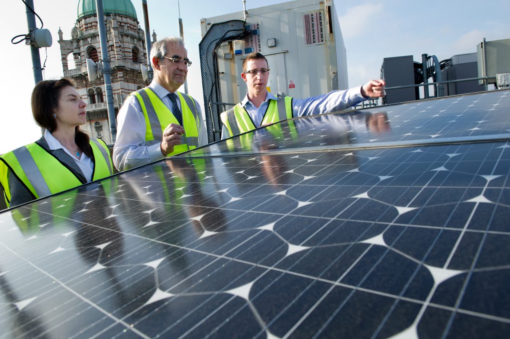 Three researchers in hi-vis vests discuss a solar panel, with the distinctive shape of Queen's Tower in the background. 