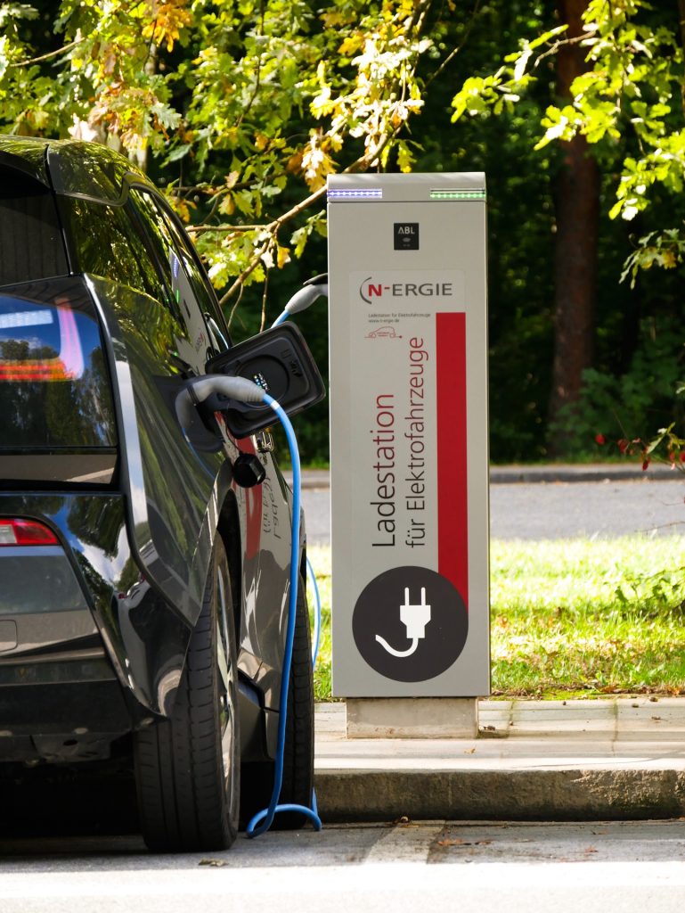 An electric car is plugged into a public charge point in a sunny carpark. 