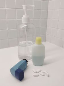 A bottle of hand sanitizer, another of shampoo, an asthma inhaler and a handful of white tablets on a bathroom shelf. 