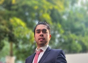 A photo of Leonardo Zea Reyes wearing a navy coloured suit with a pink tie and a background of green trees.