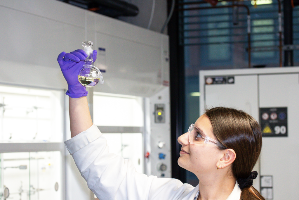Harriet Judah, wearing protective eyewear, a lab coat and purple gloves, inspecting a flask of ionic solvent.