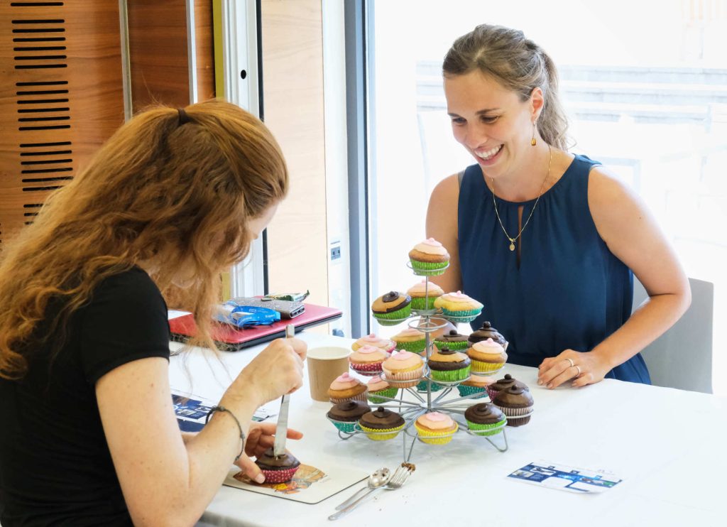 A display stand of cupcakes is in the middle of the table. Martha talks through the activity while a visitor uses a table knife to cut through the cupcake.