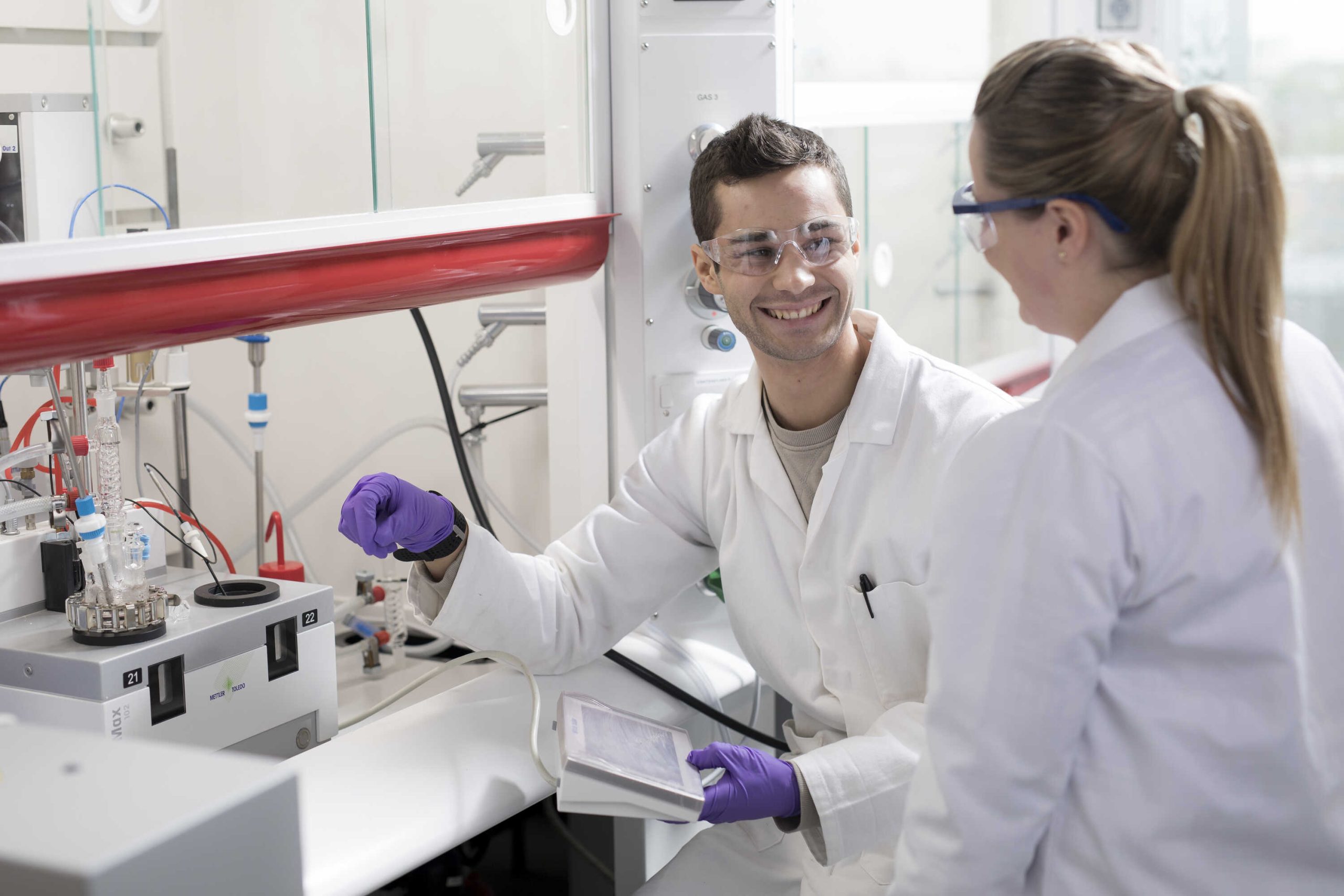 Two people wearing lab coats, gloves and goggles talking to each other in a lab