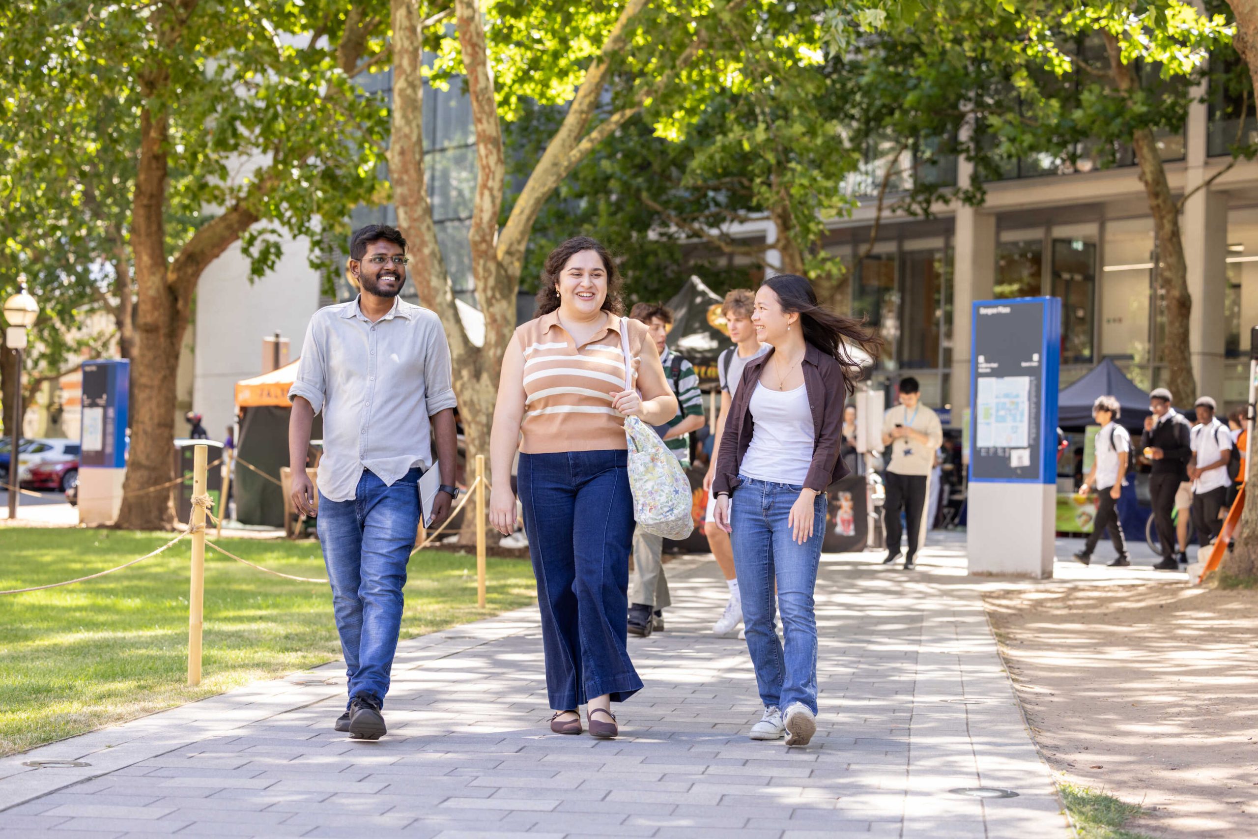 Three students walking on Imperial's South Kensington Campus, with green trees in the background and a green grass lawn next to them